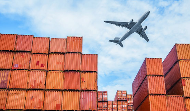 Picture of an airplane flying over a large number of stacked shipping freight containers.
