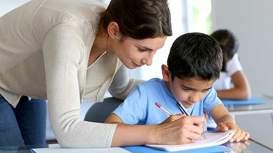 Techaer instructing child writing on a notebook and  sitting on a chair.