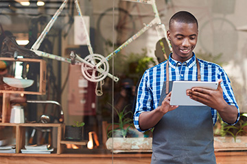 Smiling young African entrepreneur wearing an apron and standing in front of his trendy cafe working online with a digital tablet