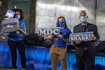 Photo of three people wearing mask protection stand at a table showing their pride for Miami Dade College by holding signs