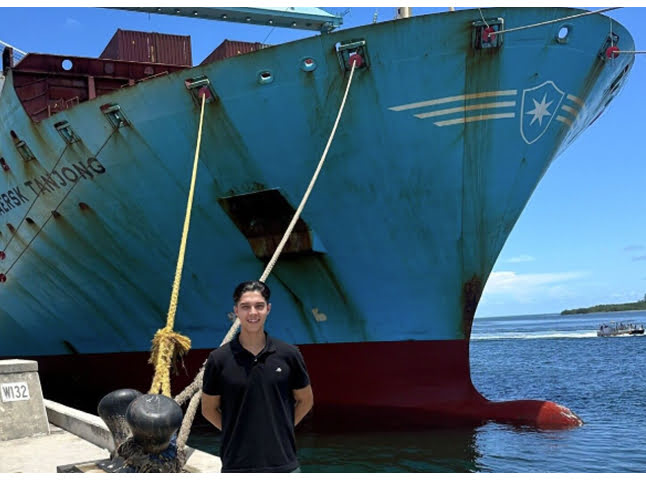 A student stands in front of a large cargo ship docked at the port.